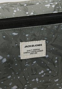 Bolsa de tela gris con un patrón moteado, cierre de cremallera negra y una etiqueta rectangular que muestra información de la marca e instrucciones de cuidado.