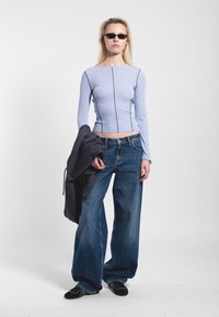 Light blue ribbed long-sleeve top with black stitching details, paired with wide-leg blue jeans, holding a dark gray sweatshirt, and black shoes.