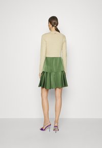 Maglione lungo a maniche con texture crema indossato sopra una gonna di seta verde con orlo a campana. La modella indossa sandali con tacco viola.