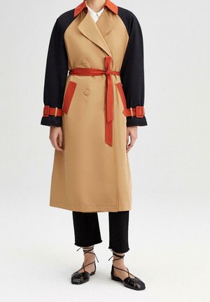Kvinde iført en beige trenchcoat med sorte ærmer, rød krave, bælte, lommedetaljer og sorte flade sko med ankremønster, stående på hvid baggrund.