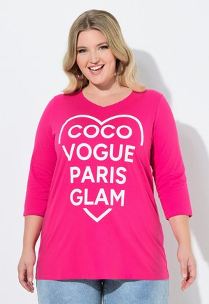 Hymyilevä vaaleatukkainen nainen pukeutuu kirkkaanpunaiseen pitkähihainen toppiin, jossa on valkoinen teksti "COCO VOGUE PARIS GLAM" sydämen ääriviivojen sisällä.