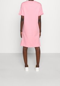 Różowa bawełniana sukienka t-shirtowa o luźnym kroju, z krótkimi rękawami, do połowy łydki, o gładkiej teksturze, noszona z białymi trampkami.