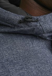 Chaqueta con capucha de tejido texturizado en azul oscuro que presenta un patrón de espiga. Cierre mediante un ojal negro y cordón en el cuello.