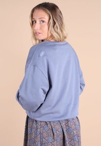 Sweat-shirt bleu clair à manches longues et poignets côtelés. Présente un petit logo en patch sur l'épaule et est associé à une jupe à motifs.