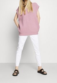 Pull en maille côtelée rose clair oversize, pantalon blanc slim-fit, et sandales sombres à double boucle, porté avec les mains dans les poches.