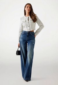 Fehér texturált, rövid fazonú dzseki gombdísszel, magas derekú, kék, bő szárú farmerrel párosítva. A modell egy kis fekete kézitáskát tart.