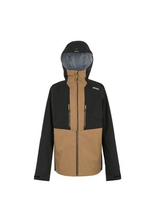 Mann trägt schwarze Beanie, schwarze gesteppte Jacke und schwarze Hose, sitzt auf weißem Block mit Hand nahe am Kopf, leicht lächelnd.