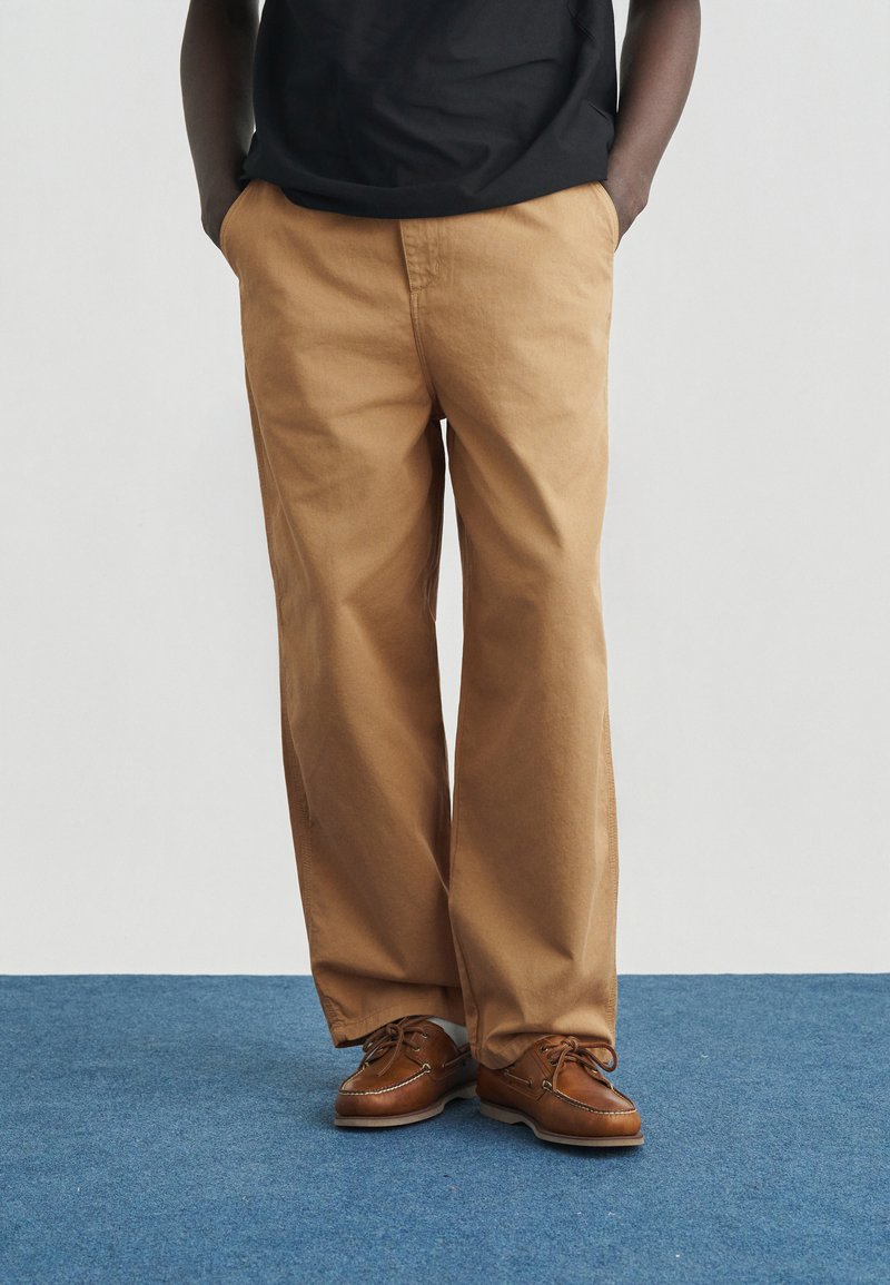 Άτομο που φοράει φαρδιά ταμπά παντελόνια, καφέ δερμάτινα παπούτσια τύπου boat, με τα χέρια στις τσέπες, στέκεται σε μπλε χαλί με λευκό φόντο.