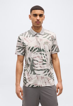 Joven con una camiseta tipo polo de manga corta con un patrón abstracto de hojas en gris, rosa y verde, y el logo EA7 en el pecho.