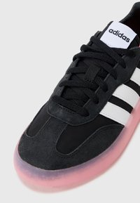 Basket en suède noir et gris avec des rayures blanches, lacets noirs et semelle translucide rose. Présente un design cousu et une languette rembourrée.