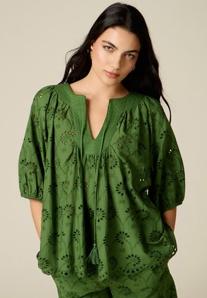 Vrouw met lang donker haar, gekleed in een losse groene geborduurde blouse met kwastjes, staand met handen in de zakken tegen een effen achtergrond.