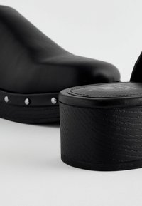 Sabot à plateforme noire avec dessus en cuir lisse, bout rond, clous argentés décoratifs, et semelle en bois texturée sur un fond neutre.