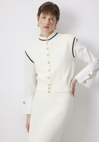 Witte gebreide vest met zwarte rand, gouden knoopsluitingen en geribde onderkant, gelaagd over een witte lange mouw shirt. Gladtextuur, getailleerd ontwerp.
