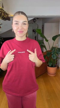 Rotes Baumwoll-T-Shirt mit kurzen Ärmeln, versehen mit dem "PEGA-DOR"-Logo. Das Modell zeigt auf das T-Shirt und steht in einem Raum mit Pflanzen.