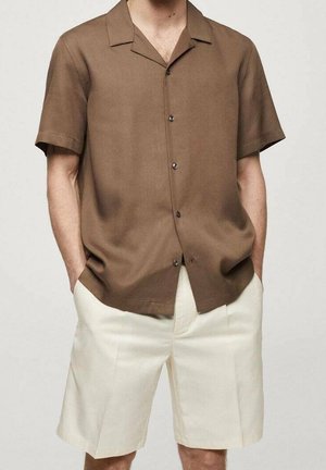 Mężczyzna w brązowej koszuli z krótkim rękawem na guziki i kremowych krótkich spodenkach do kolan, z rękami w kieszeniach.