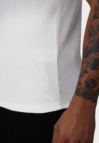 Weißes Sportshirt aus strukturiertem, geripptem Stoff. Es hat ein dezentes Logo an der Seite und einen abgerundeten Saum. Wird mit schwarzen Unterteilen getragen.