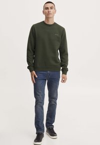 Donkergroene sweatshirt met ronde hals en geribbelde boorden, gecombineerd met blauwe spijkerbroek en zwarte sneakers. Eenvoudige logodetail op de borst.