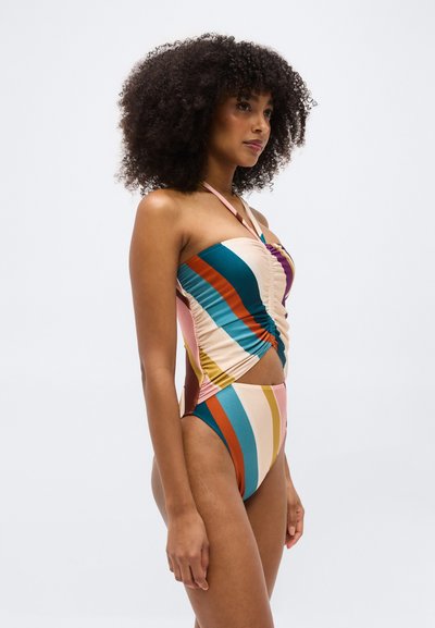 Γυναίκα με κατσαρά μαλλιά που φοράει εφαρμοστό πολύχρωμο ριγωτό μαγιό χαλτέρ, στέκεται προφίλ μπροστά σε απλό φόντο.