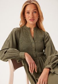 Robe longue à manches longues vert olive avec un bustier smocké, des boutons sur le devant et des manches texturées. Tissu doux, col élégant et coupe décontractée.