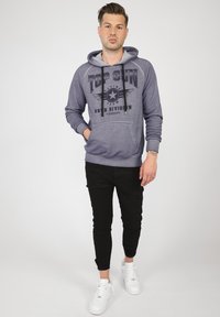 Grå huvtröja med "TOP GUN"-grafik, snörning och framficka. Kombineras med svarta slimmade byxor och vita sneakers.