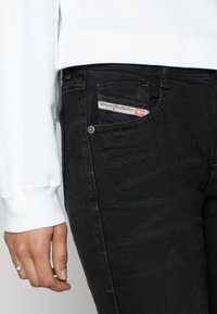 Acercamiento a una persona que lleva puesto un pantalón Diesel negro con la etiqueta del bolsillo delantero visible y una camisa blanca de manga larga con un puño y una mano anillada.