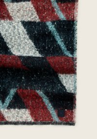 Couverture en laine avec un motif géométrique dans des couleurs rouge, noir, blanc et bleu clair, texture douce, et une étiquette noire dans le coin.