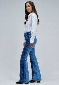 Mujer de pie de lado, vistiendo una camisa blanca de manga larga, jeans acampanados bicolor en tonos azules y zapatos de tacón negros, contra un fondo liso.