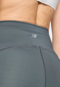 Leggings gris taille haute en tissu extensible avec des coutures plates et un petit logo New Balance blanc sur la ceinture.