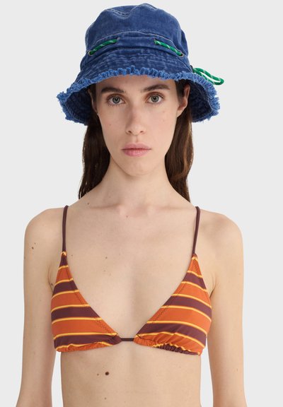 Jeune femme portant un chapeau seau bleu avec des détails verts et un haut de bikini rayé orange et marron, face à l'avant sur un fond uni.