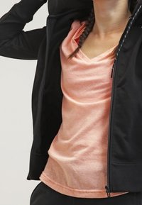 Un sweat à zip noir porté sur un t-shirt à col en V rose clair. Le sweat présente un tissu lisse et des poignet côtelés, tandis que le t-shirt est doux avec une légère texture.