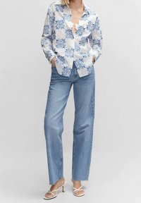 Camisa blanca con patrón floral azul, por fuera del pantalón, combinada con jeans rectos de un azul claro y tacones blancos con tiras. Textura de tela suave.