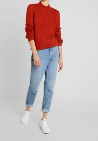 Červený pletený svetr s ribované texturou, zkombinovaný se světle modrými zúženými džíny a bílými teniskami. Svetr má vysoký límec.