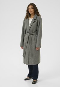 Pelēks garš mētelis no mīksta vilnas maisījuma, ar iebūvētu atloku, jostu viduklī, divām priekšējām kabatām un brīvu piegriezumu virs džinsa biksēm.