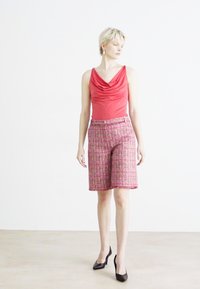 Czerwony top z dekoltem w łódkę i bez rękawów, zestawiony z różowymi, wzorowanymi szortami o długości do kolan. Modelka ma na sobie czarne, wysokie obcasy. Proste tło.