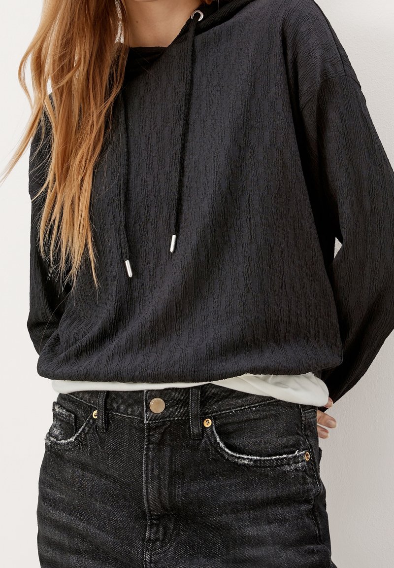 Sudadera negra texturizada con cordón y extremos en plateado. Combinada con unos jeans negros de cintura alta con detalles desgastados.