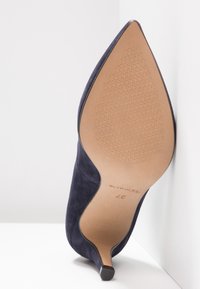 Chaussure à talon haut pointu en daim bleu marine avec une semelle texturée et un petit talon aiguille, inclinée sur un fond blanc. Étiquette de taille 37 visible.