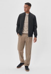 Must mustritsukas ülerõivas, beež lukuga kampsun, beež püksid ja mustad tossud pruunide talladega, poseerivad lihtsa valge tausta ees.