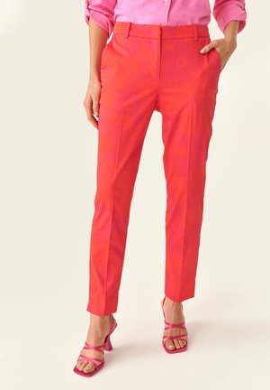 Pantalons à motif floral rouge avec une coupe fuselée, fabriqués à partir de tissu lisse. Portés avec des talons roses à lanières et une chemise boutonnière rose pâle.