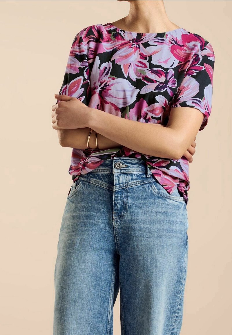 Blommig blus med kort ärm i rosa och lila mönster på en svart bakgrund. Matchas med ljusblå vida jeans.