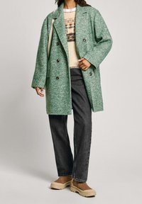 Green herringbone-patterned coat with a loose fit, double-breasted front, large buttons, paired with a beige sweater and dark jeans.