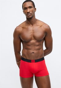 Sarkanas bokseršortenes no elastīga auduma ar melnu elastīgu jostu, uz kuras ir redzams logotips. Produkts ir pieguļošs, izceļot ķermeņa kontūras. Minimālistisks dizains.