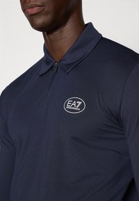 Mornarsko polo srajca z teksturirano tkanino, kratkim ovratnikom in delnim zadrgo. Ima belo EA7 logo na zgornjem levem prsnem delu.
