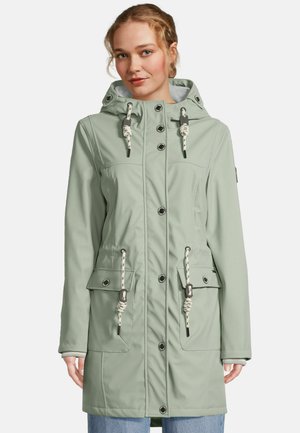 Femme portant un imperméable à capuche vert clair avec boutons et cordons de serrage, debout devant un fond uni.