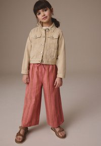 Lichtbeige spijkerjack met twee voorzakken, gecombineerd met roze wijde broek met verticale strepen en bruine sandalen.