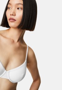 Weißer Bra mit dünnen Trägern, glattem Stoff und einem dezenten Schimmer. Besitzt eine konturierte Form mit einer zarten Borde entlang der Kanten.