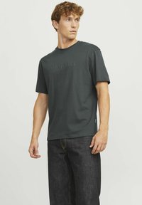 Tumši pelēks t-krekls ar īsām piedurknēm, ar teksturētu "ORIGINALS" uzrakstu priekšpusē. Apvienots ar tumši zilu džinsu biksēm.