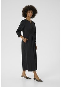 Black cotton dress with a relaxed fit, pleated bodice, 3/4 sleeves, side pockets, and knee-length hem, paired with striped kitten heels.