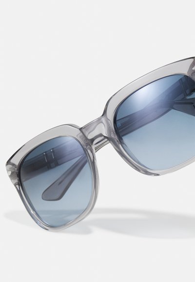 Óculos de sol oversized cinza transparente com lentes degradê azuis e hastes retas. Apresenta um design elegante e moderno com um acabamento brilhante.