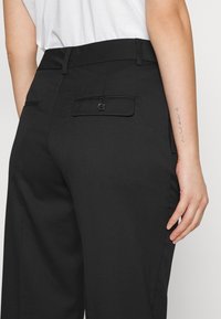 Calças pretas com uma textura suave, design de perna reta e bolsos traseiros com fechos de botão. Exibidas ao lado de um braço com uma tatuagem.