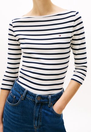 Frau trägt ein weißes und marineblau gestreiftes Bootshals-Oberteil mit dreiviertel Ärmeln, das in eine hoch taillierte blaue Jeans aus Denim gesteckt ist.